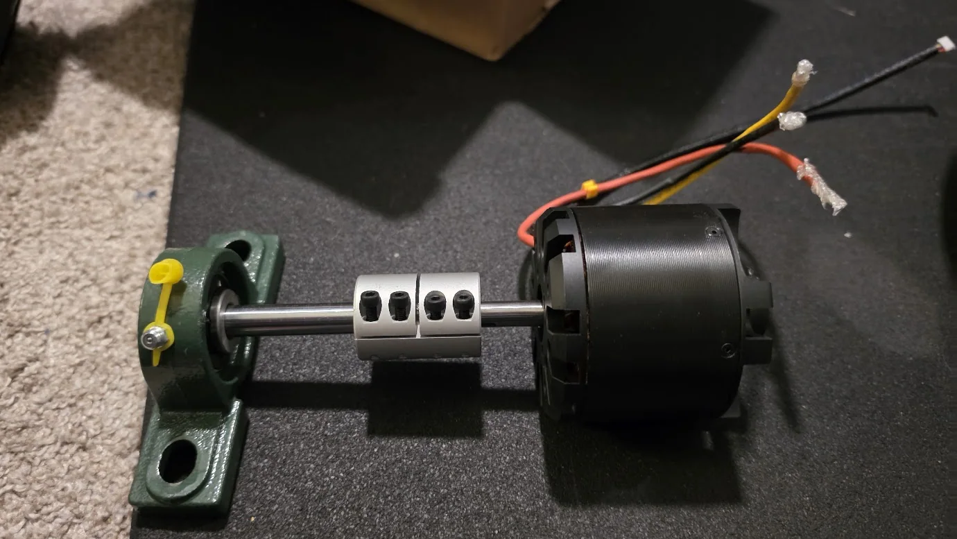 Photo of the first real parts on a workbench: the black brushless motor, a silver clamp coupling and a 12 mm steel shaft running into a bearing